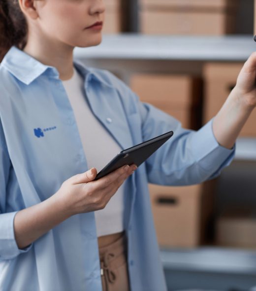 Close-up of young woman in blue shirt scanning qr codes on packed boxes with ordered online goods standing on shelves in storage room